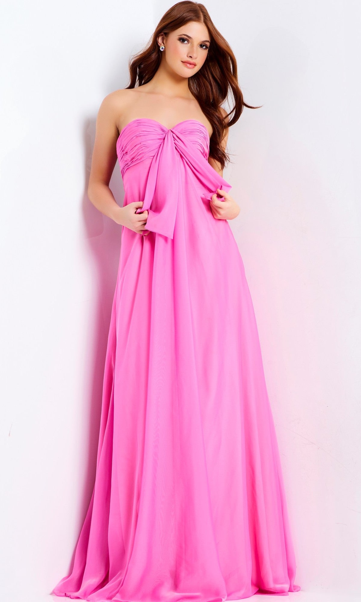A woman stands against a white background wearing Jovani 47041, a long strapless chiffon prom dress in bright pink with a large chest bow.